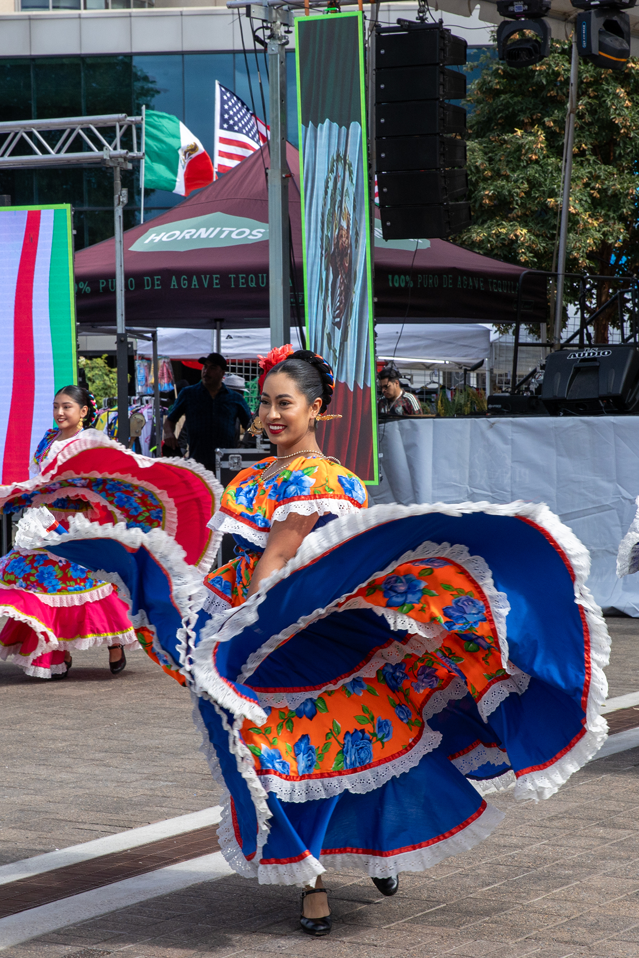 A group of dancers performs traditional Mexican folkloric dance at El Grito Portland 2024. The lead dancer smiles brightly while twirling a colorful blue and orange dress with floral patterns and lace trim, showcasing cultural pride and movement onstage.