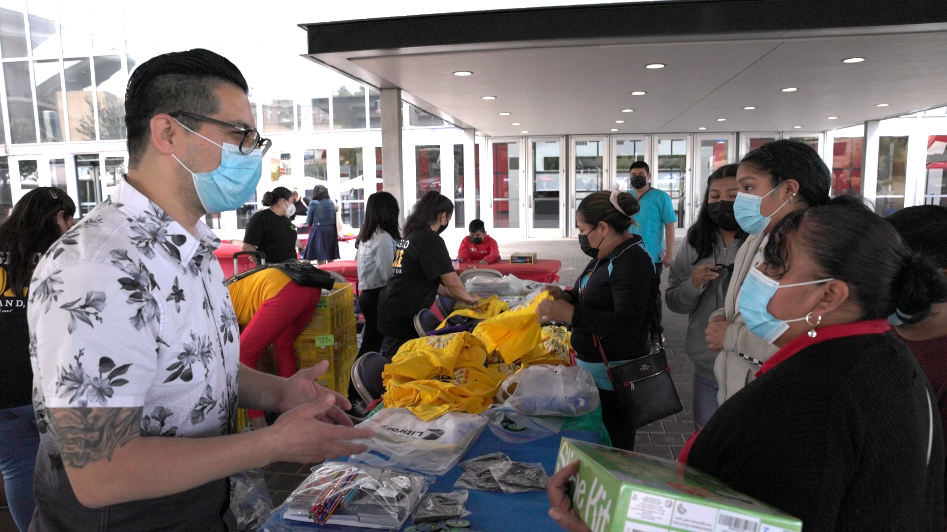 A community outreach representative speaks with a group of attendees at an informational booth during a El Grito. The table is filled with resources, flyers, giveaways, and educational resources, while people browse and engage with the staff.
