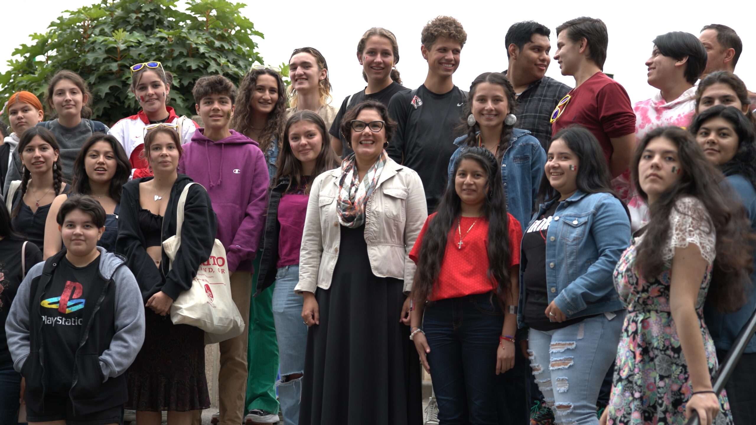 A group of high school students from Portland Public Schools stands with a local government leader; during the school field trip at El Grito Portland. The students are smiling and gathered outdoors, many with festive face paint and casual attire.