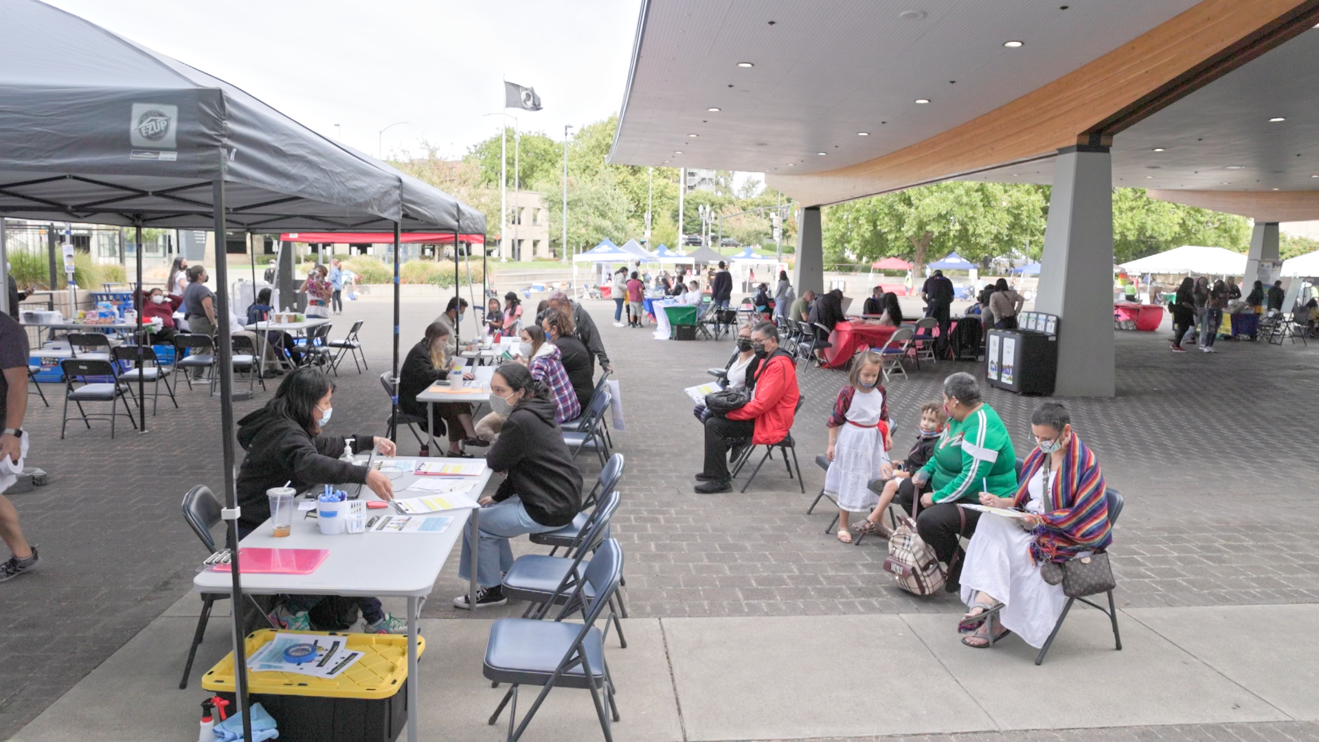 Community members gather at El Grito Portland's health services area featuring multiple booths under canopies. People sit at tables or wait in line to receive COVID-19 vaccinations, flu shots, glucose testing, and cholesterol screenings.