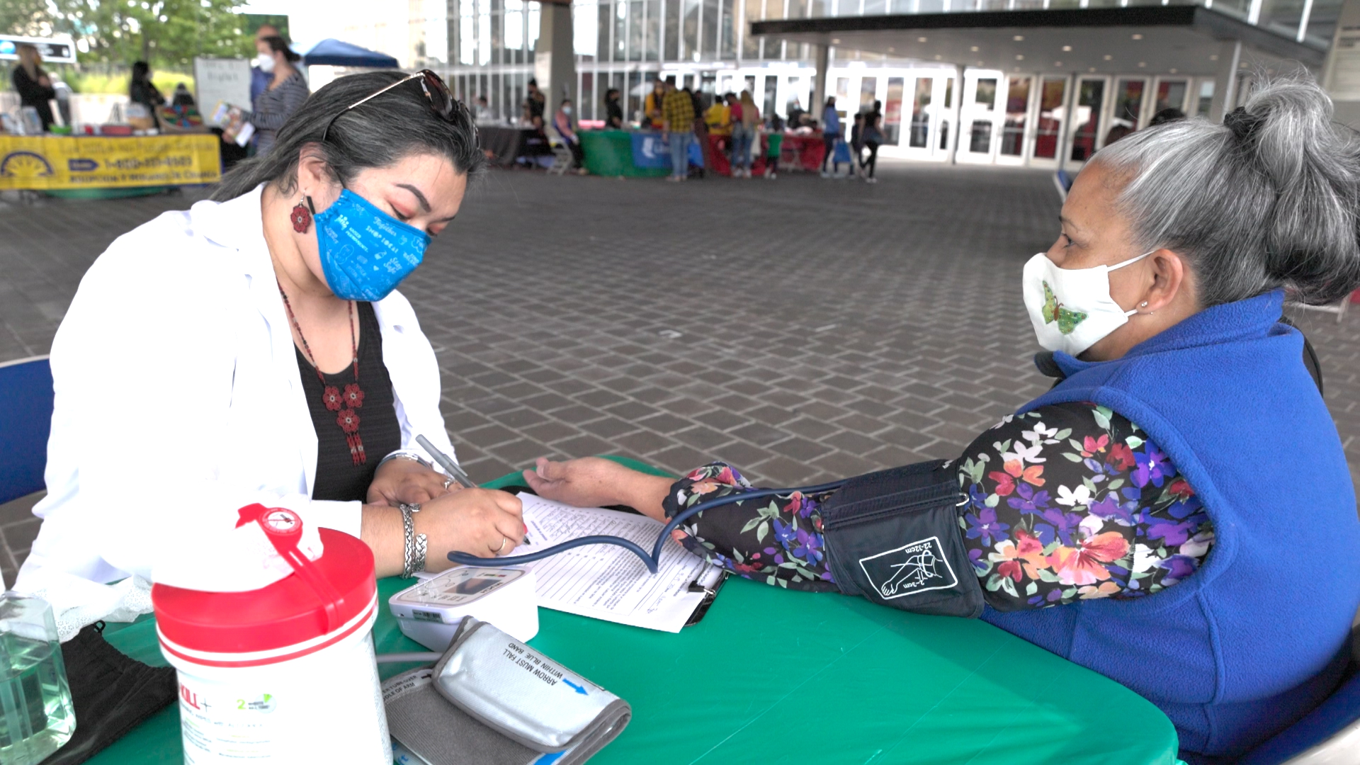 A smiling woman browses traditional clothing at a vendor booth during El Grito Portland. She holds a white embroidered garment while colorful sombreros and Mexican paper rosettes hang above. Other attendees explore the cultural market in the background.
