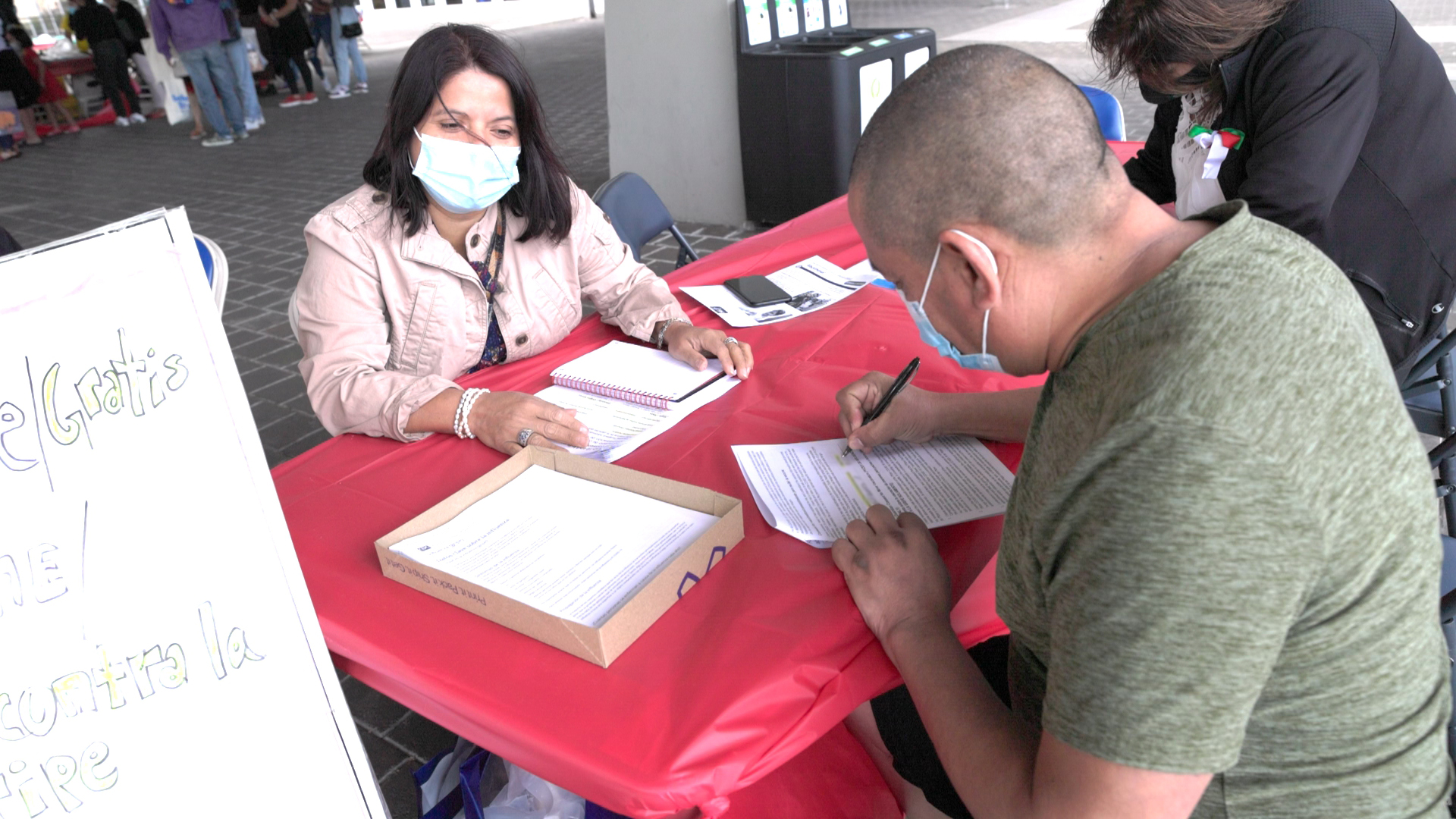 A man fills out a vaccination consent form at a health resource booth during El Grito Portland. A staff member sits across from him at a red-covered table with paperwork and informational materials..