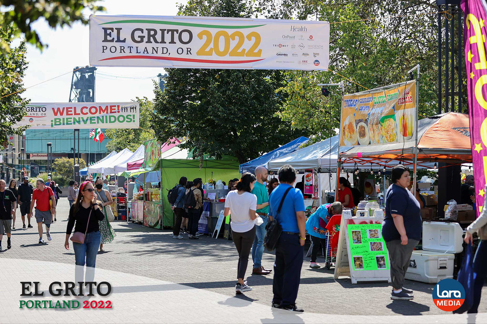 People walk through a vibrant outdoor food court at El Grito Portland. Overhead banners read “EL GRITO PORTLAND, OR 2022” and “WELCOME! ¡BIENVENIDOS!” Food booths display menus featuring aguas frescas and tacos, while attendees browse, eat, and enjoy the festival atmosphere.