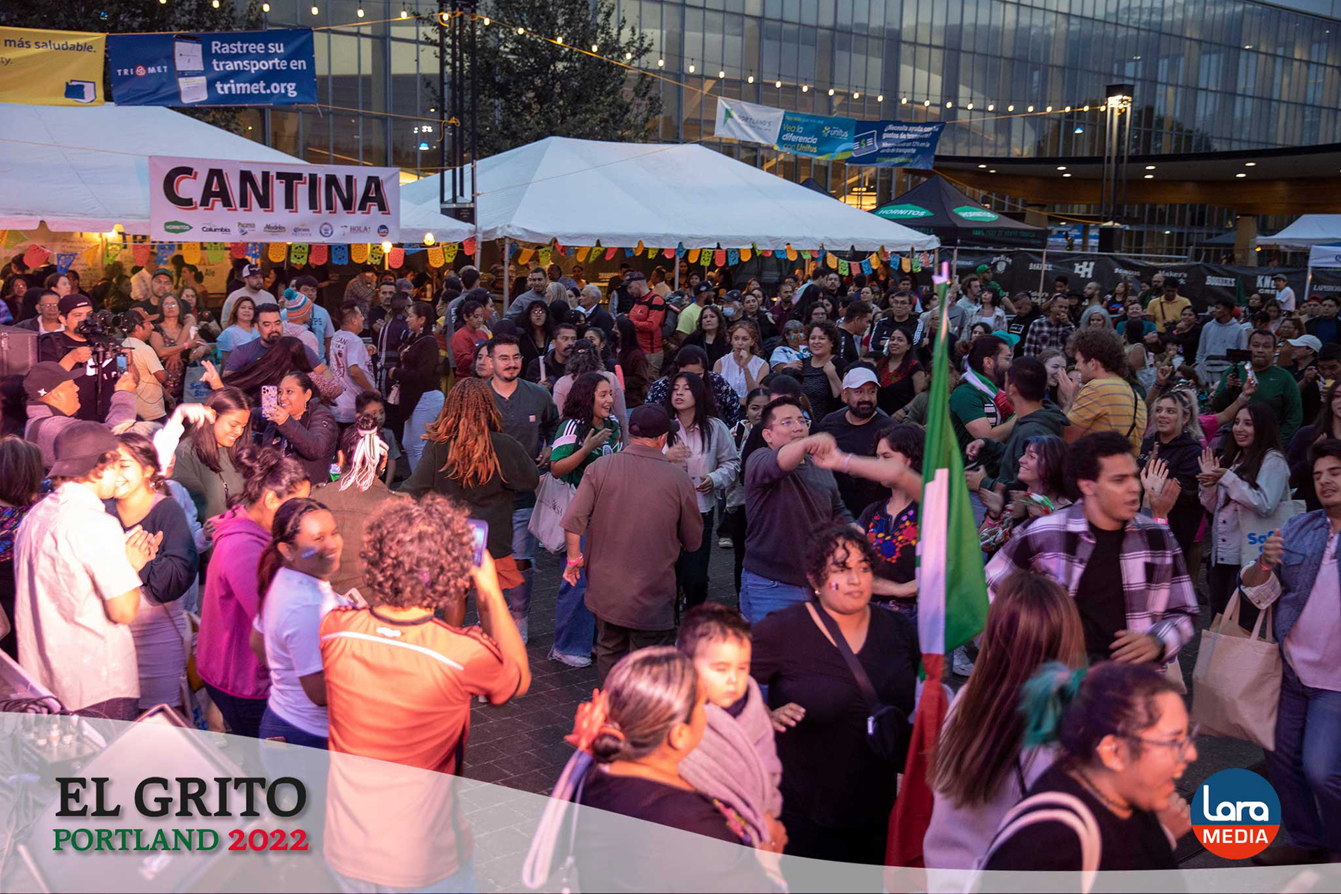 A large crowd dances and celebrates in front of the stage at El Grito Portland. The festive evening scene is filled with colorful lights, music, food vendors, and hundreds of people of all ages enjoying the event.