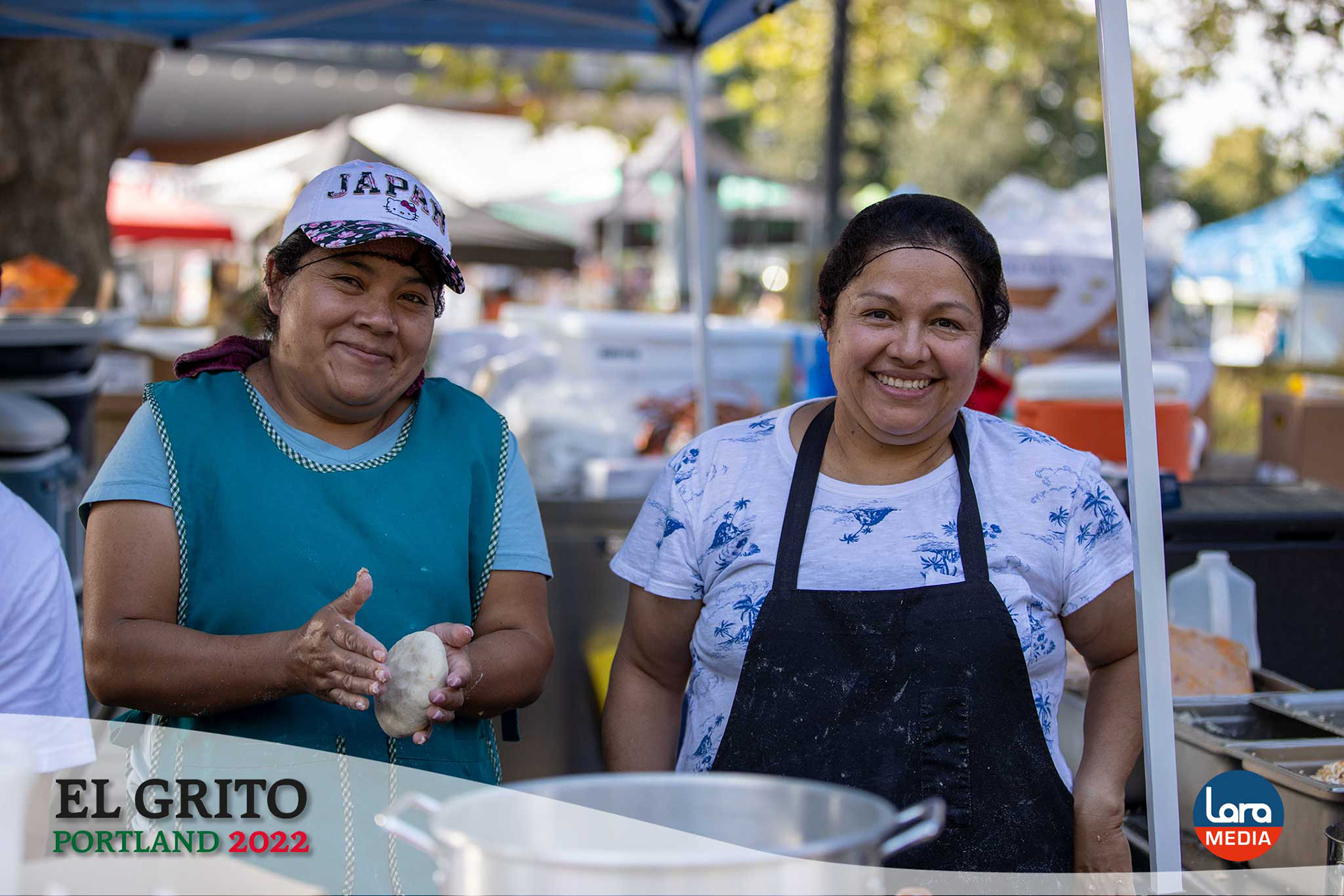 Two women smile while preparing fresh food at their booth during El Grito Portland. One forms dough with her hands, while the other stands beside her wearing a kitchen apron. They are surrounded by cooking equipment under a vendor tent at the outdoor festival.