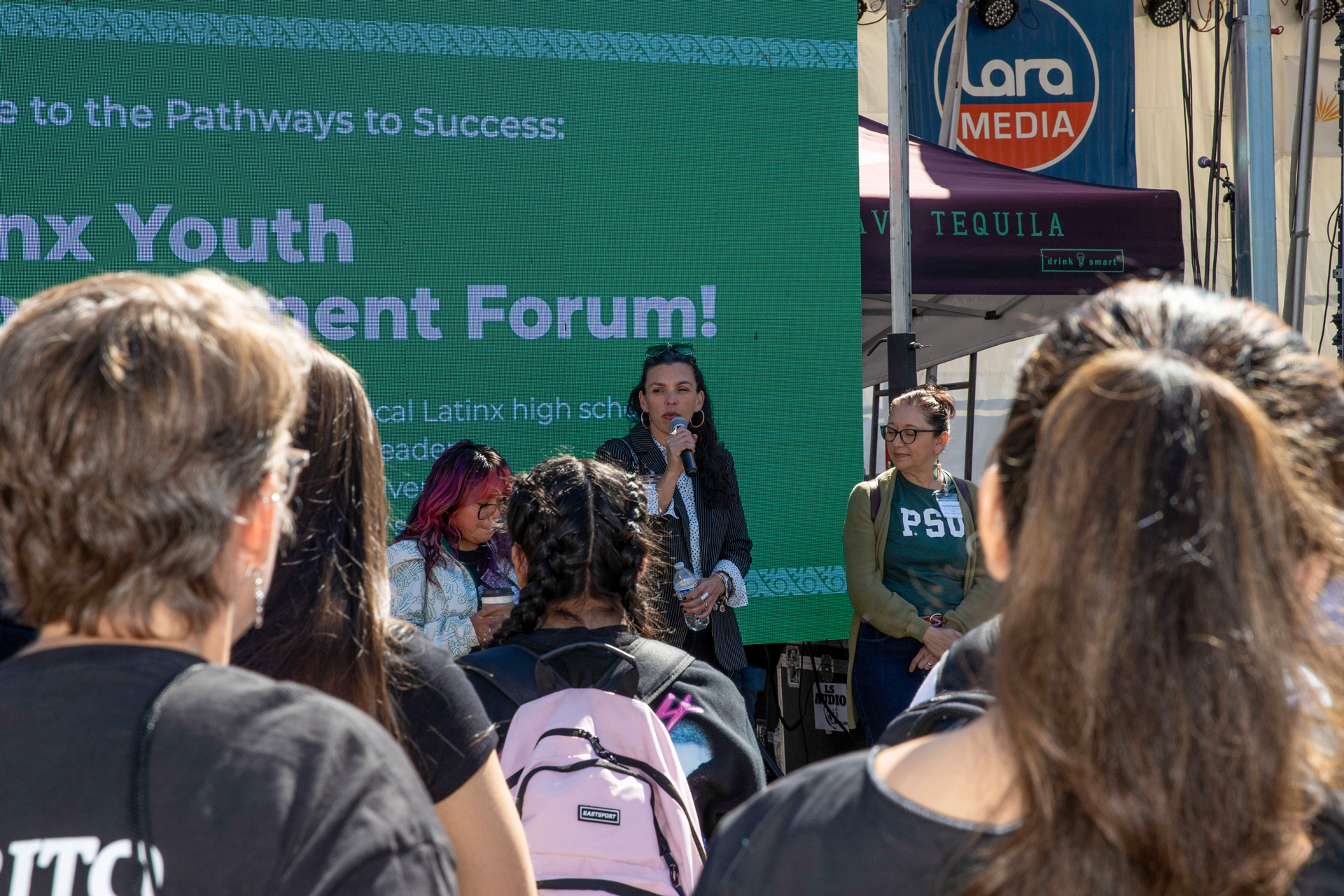 A smiling woman browses traditional clothing at a vendor booth during El Grito Portland. She holds a white embroidered garment while colorful sombreros and Mexican paper rosettes hang above. Other attendees explore the cultural market in the background.