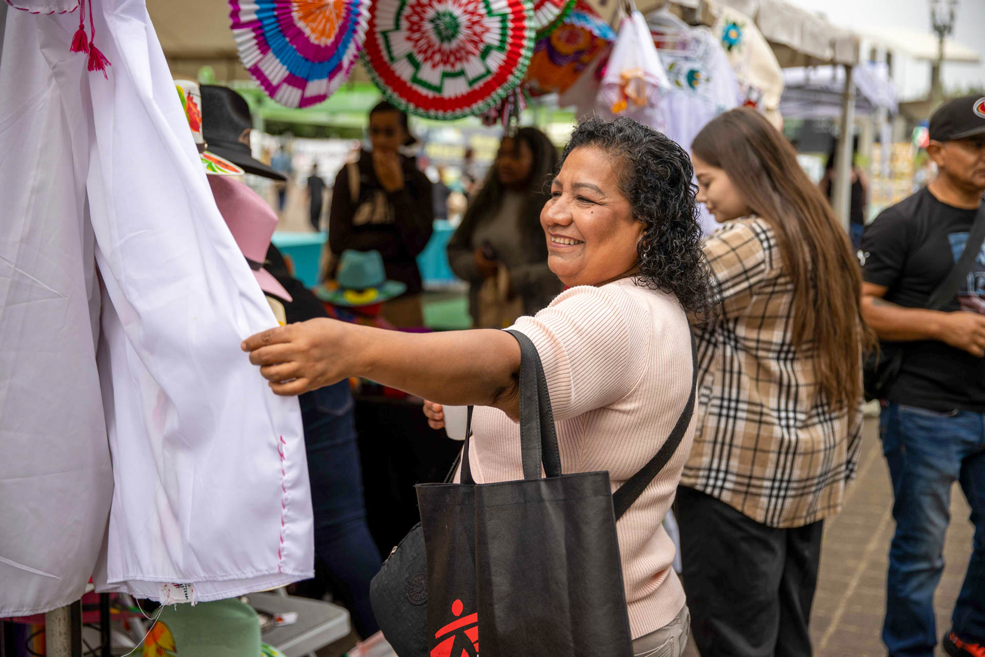 A smiling woman browses traditional clothing at a vendor booth during El Grito Portland. She holds a white embroidered garment while colorful sombreros and Mexican paper rosettes hang above. Other attendees explore the cultural market in the background.