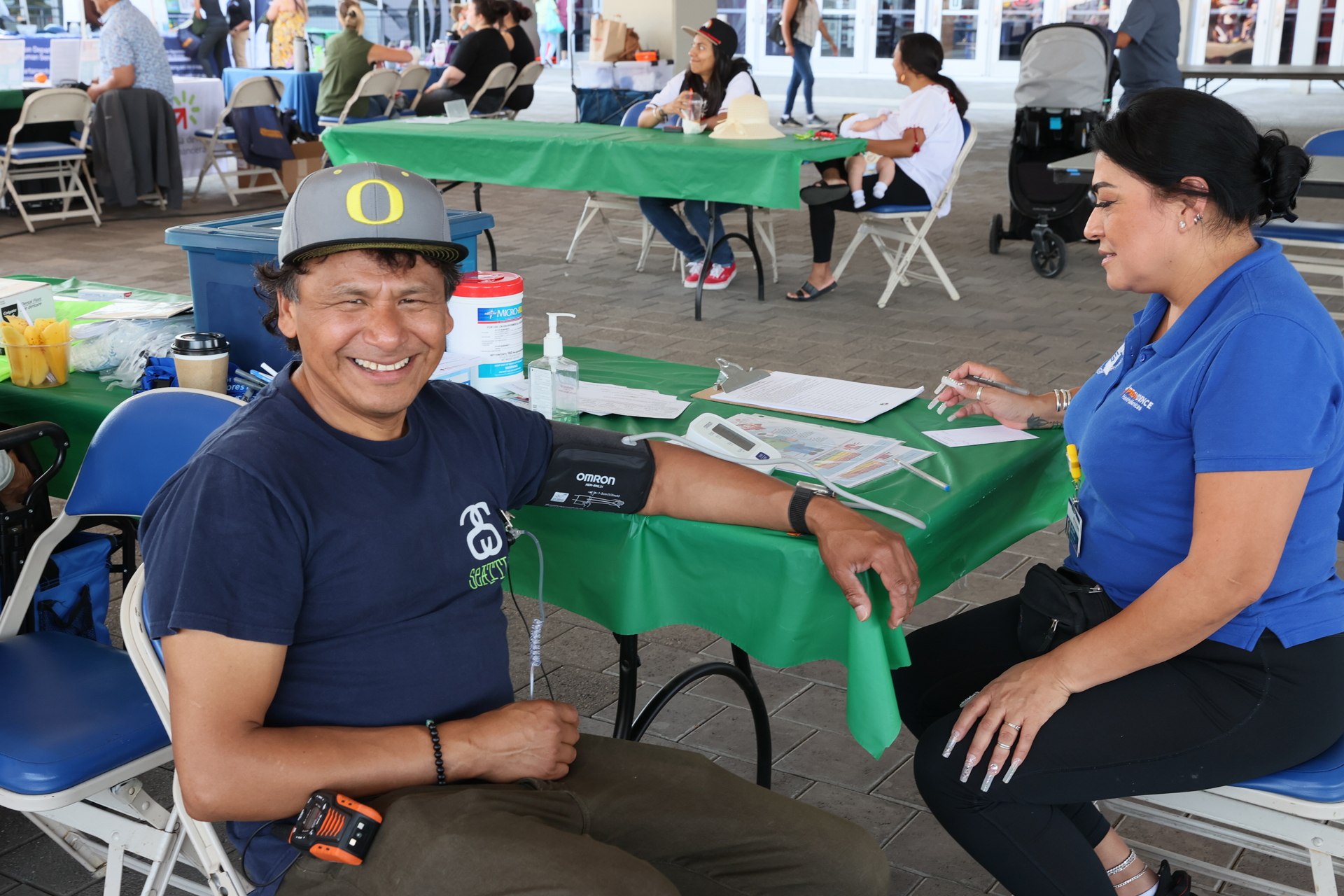 A smiling man sits at a health screening booth while a healthcare worker records his blood pressure results. They are located in the health partners area, surrounded by informational materials.