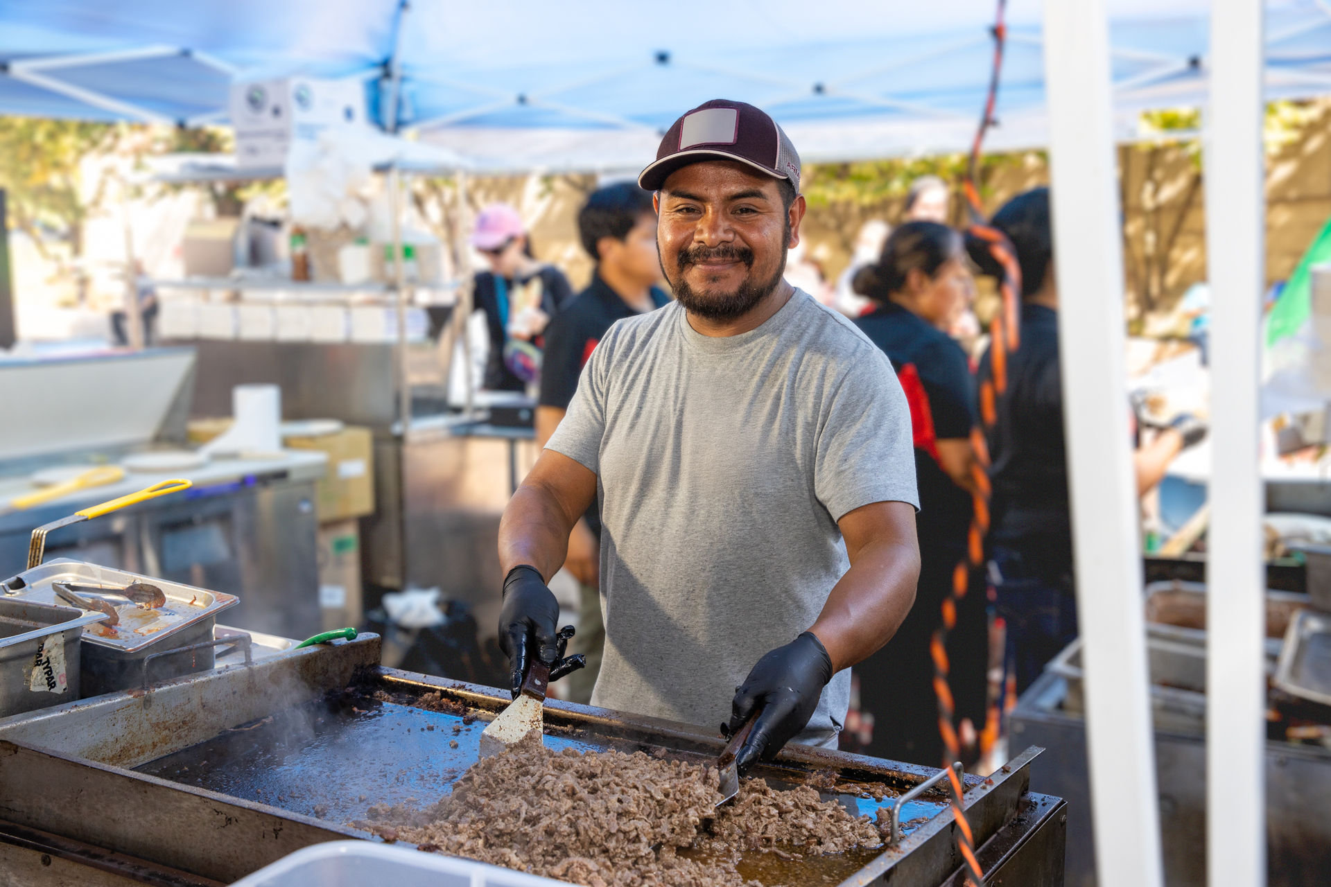 A smiling woman browses traditional clothing at a vendor booth during El Grito Portland. She holds a white embroidered garment while colorful sombreros and Mexican paper rosettes hang above. Other attendees explore the cultural market in the background.