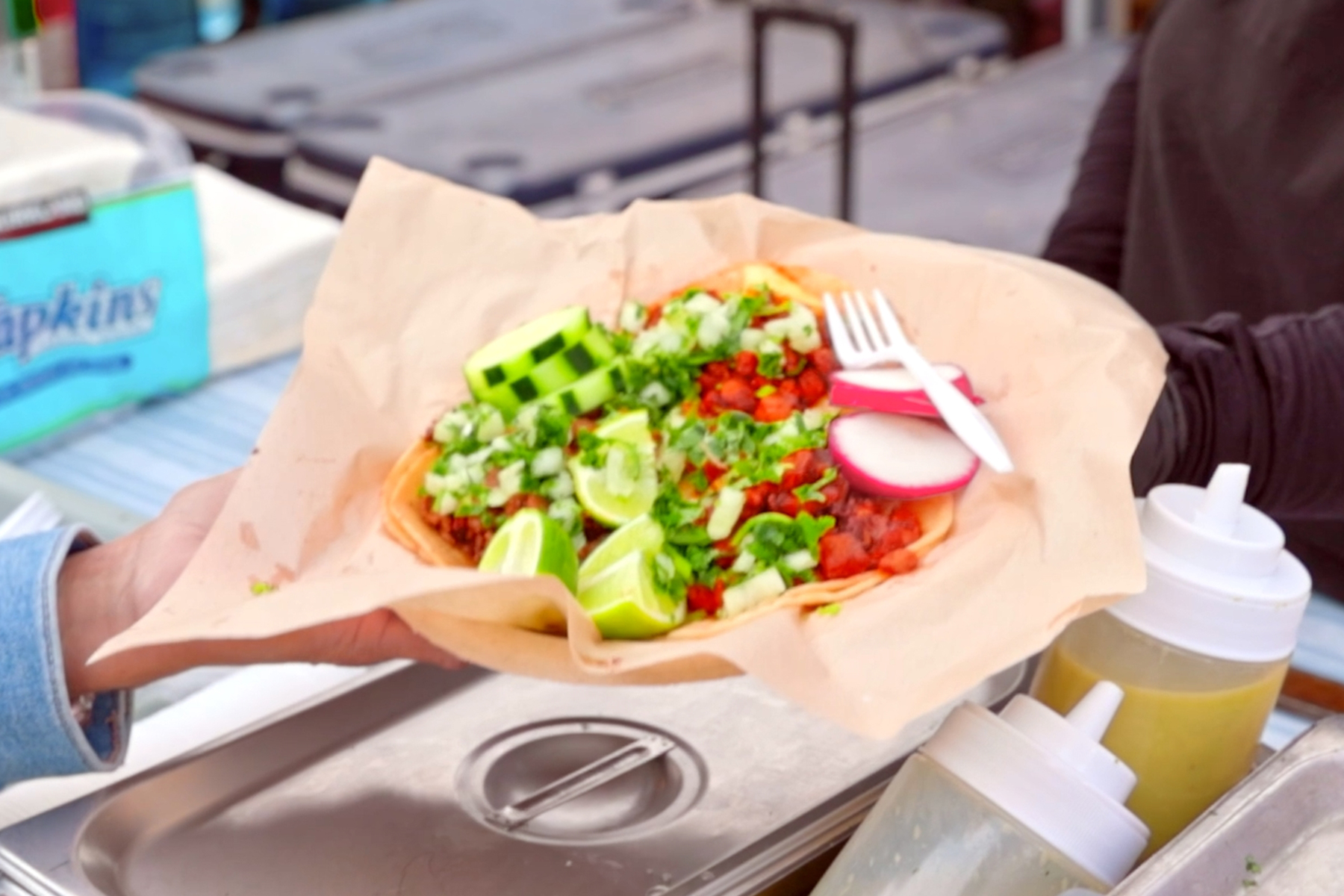 A food vendor hands a customer traditional mexican tacos topped with fresh radish, onion, and cilantro at an outdoor food court.
