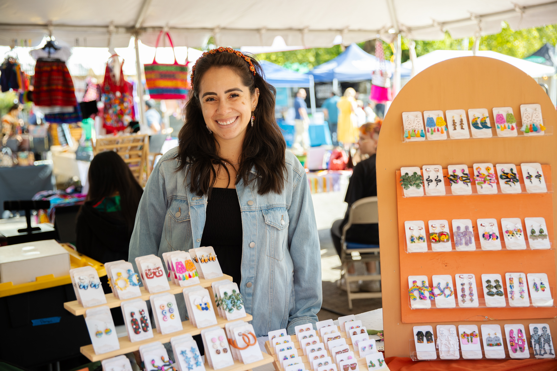 A woman entrepreneur smiles proudly behind her market booth, which is filled with displays of her colorful, handmade earrings and jewelry.
