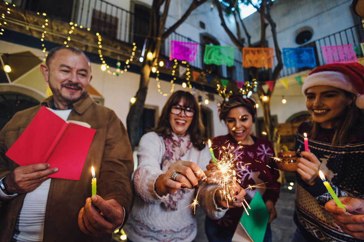 A smiling family holds candles and sparklers in a festive courtyard, celebrating Las Posadas at night under string lights and colorful *papel picado* banners.